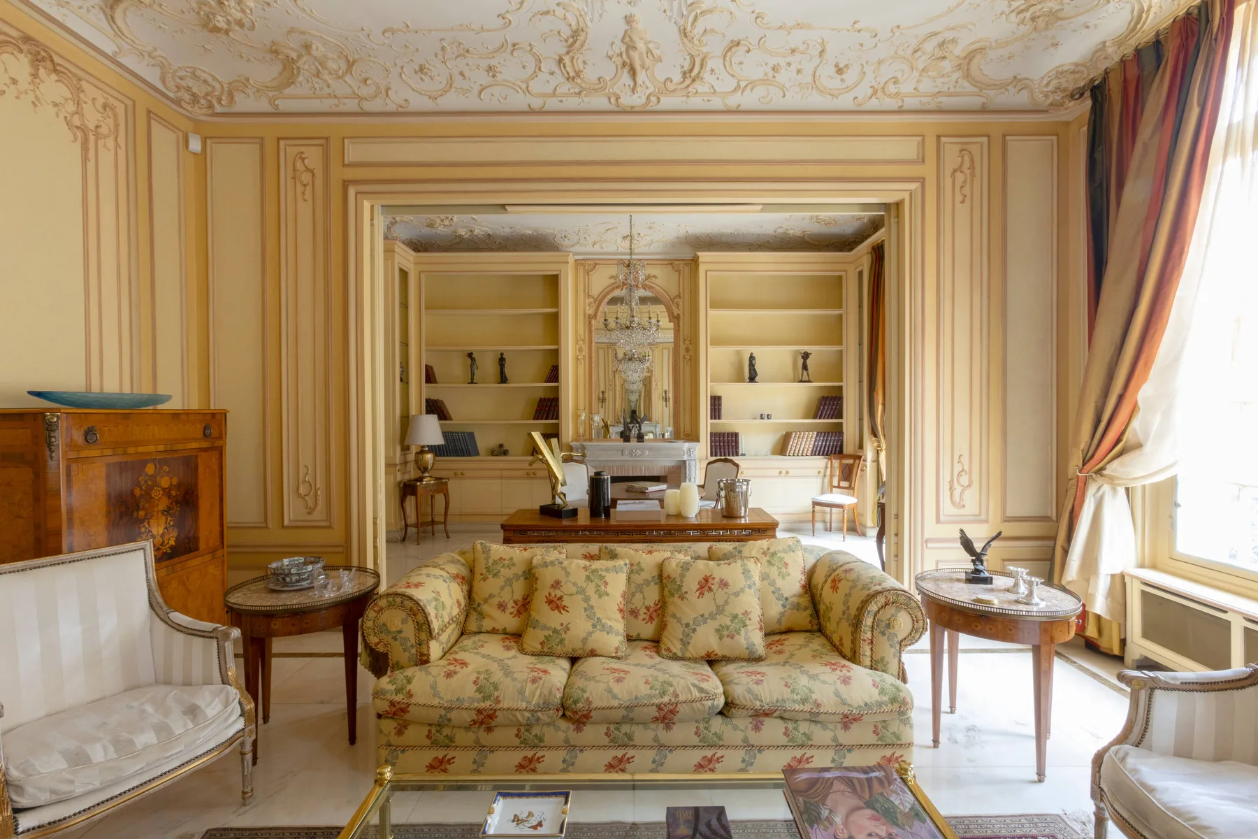 An ornate sitting room with patterned seating, built-in shelves, and tall draped windows.
