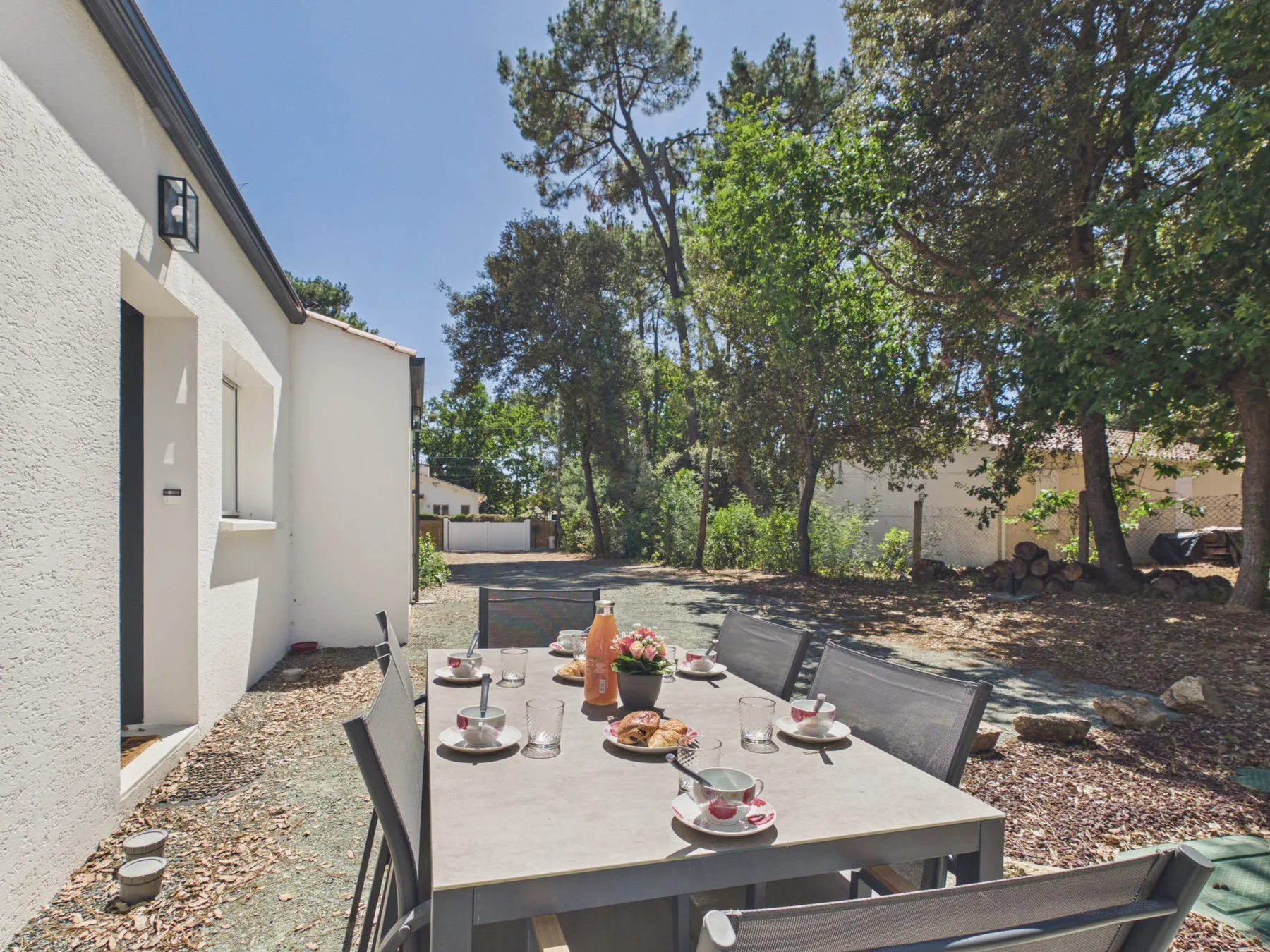 An outdoor dining area beside a white house, shaded by leafy trees.