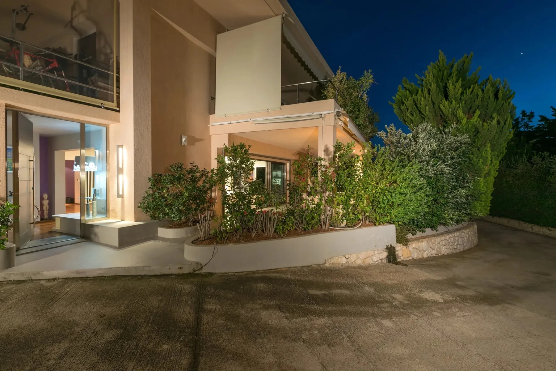 Exterior de una villa por la noche, con una entrada iluminada y vegetación alrededor.