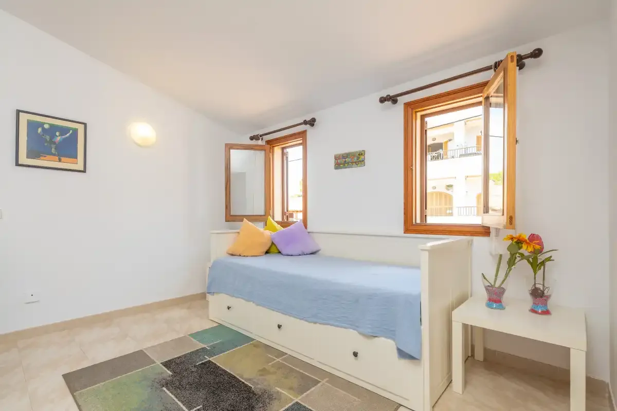 A bedroom with a bed beneath two open windows and a small bedside table.