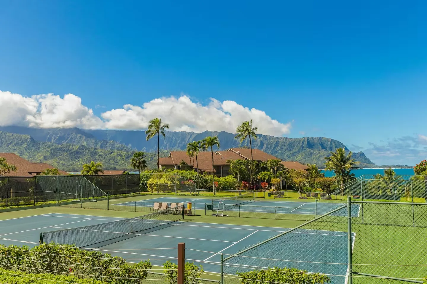 Outdoor tennis courts sit beside green lawns, with palm trees and mountains in the background.