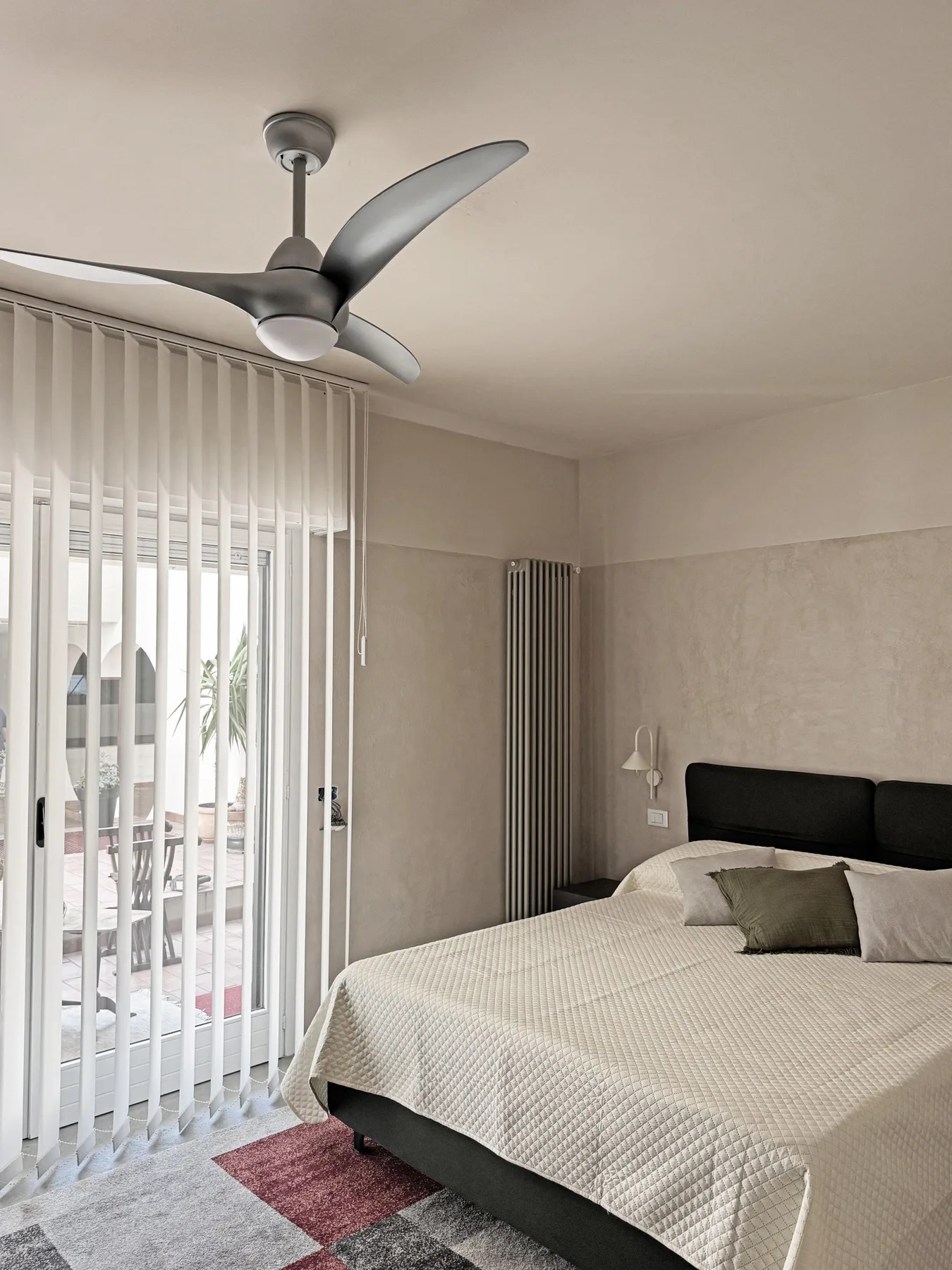 Un dormitorio con una cama junto a ventanas altas con persianas verticales.