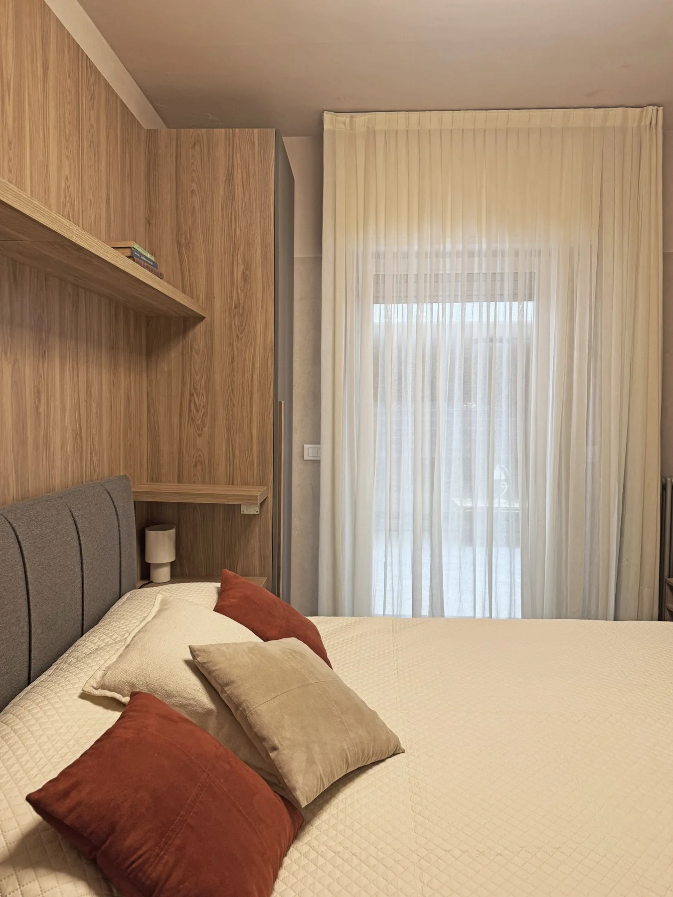 Un dormitorio con una cama, varios cojines y cortinas translúcidas en la ventana.