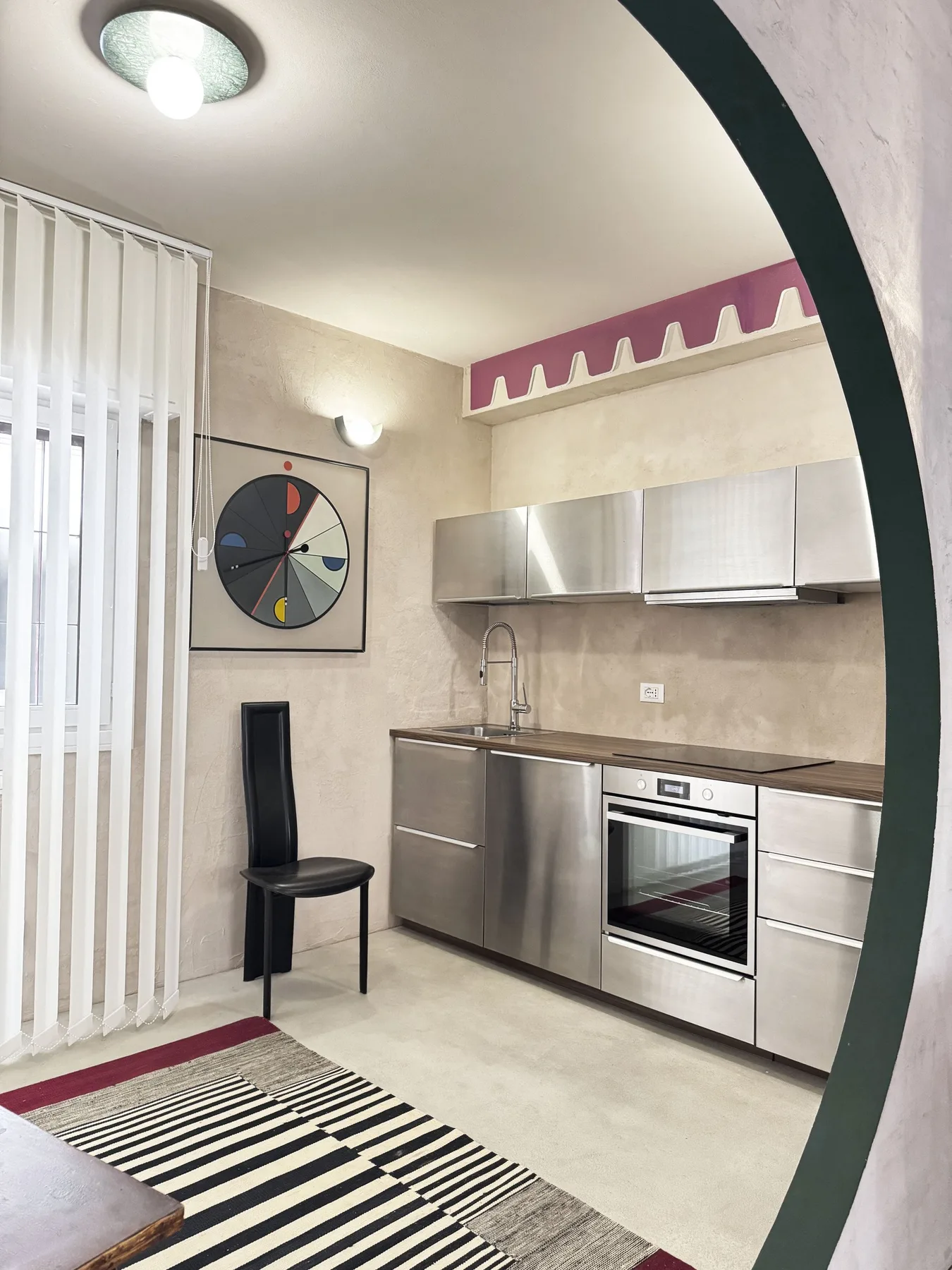 Una cocina compacta con armarios de acero inoxidable y una alfombra a rayas junto a la ventana.