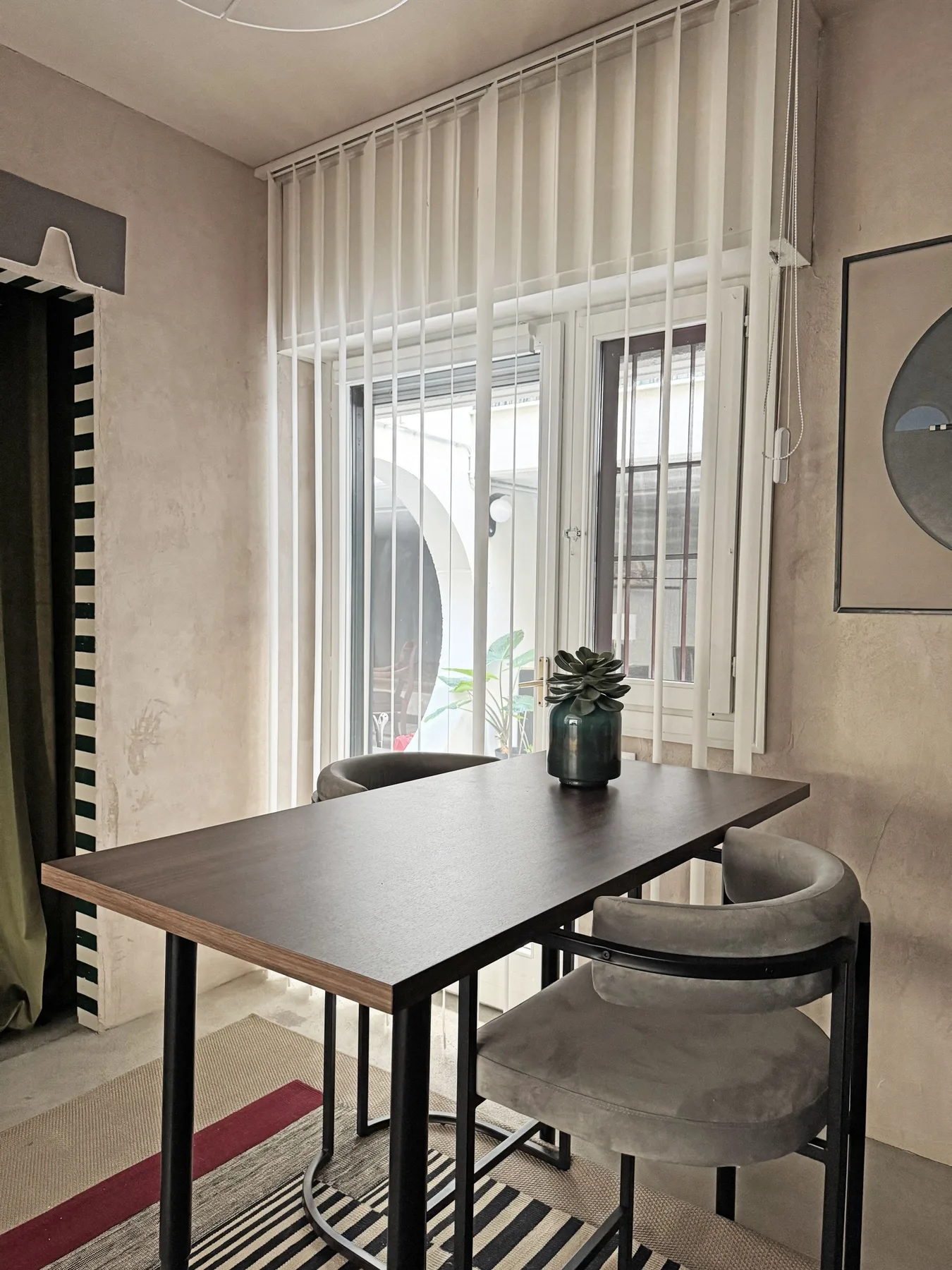 Una pequeña zona de comedor con una mesa oscura, asientos tapizados y ventanas altas.