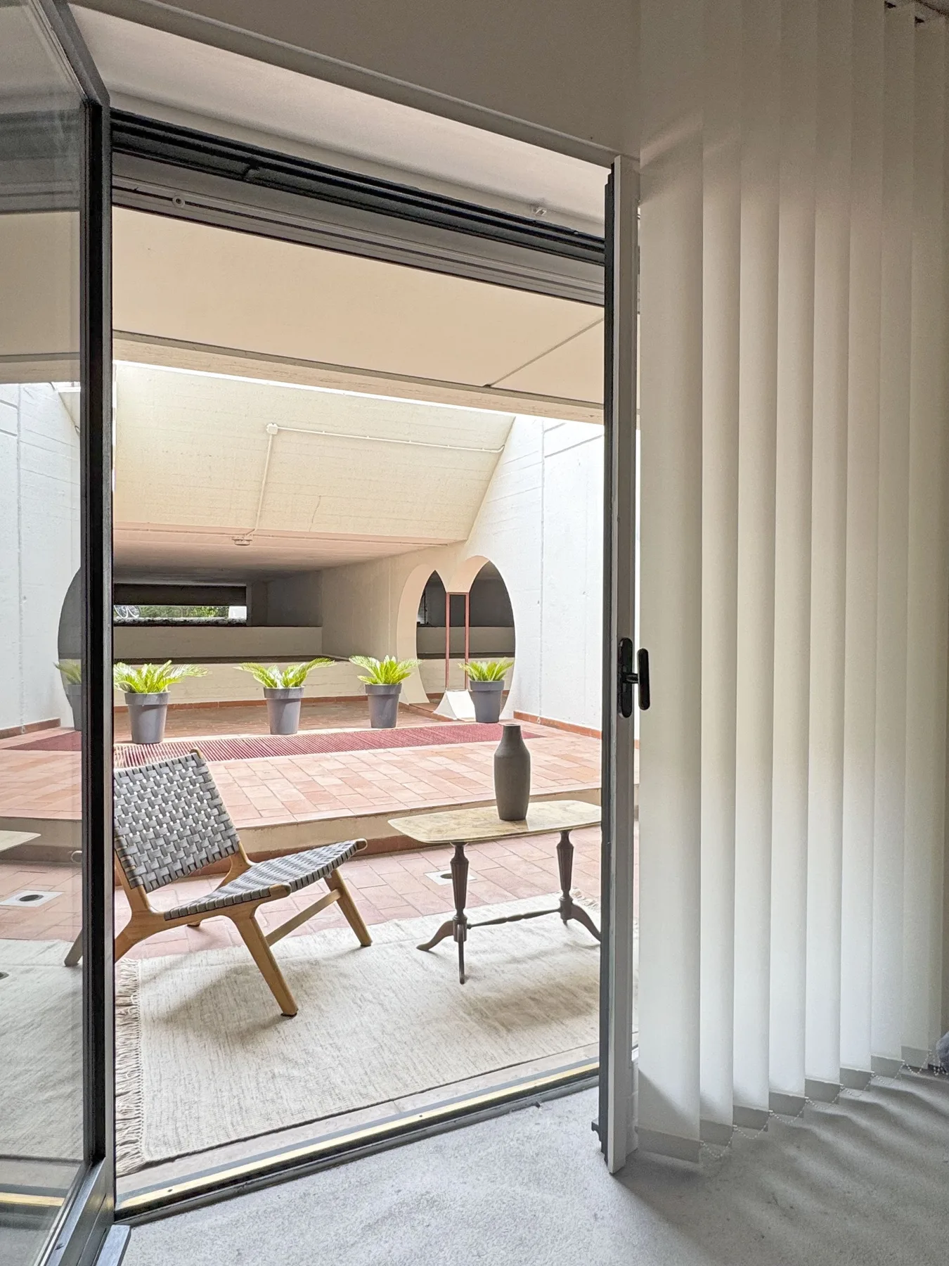Un patio luminoso se ve a través de una puerta corrediza abierta, con una silla y una mesa.