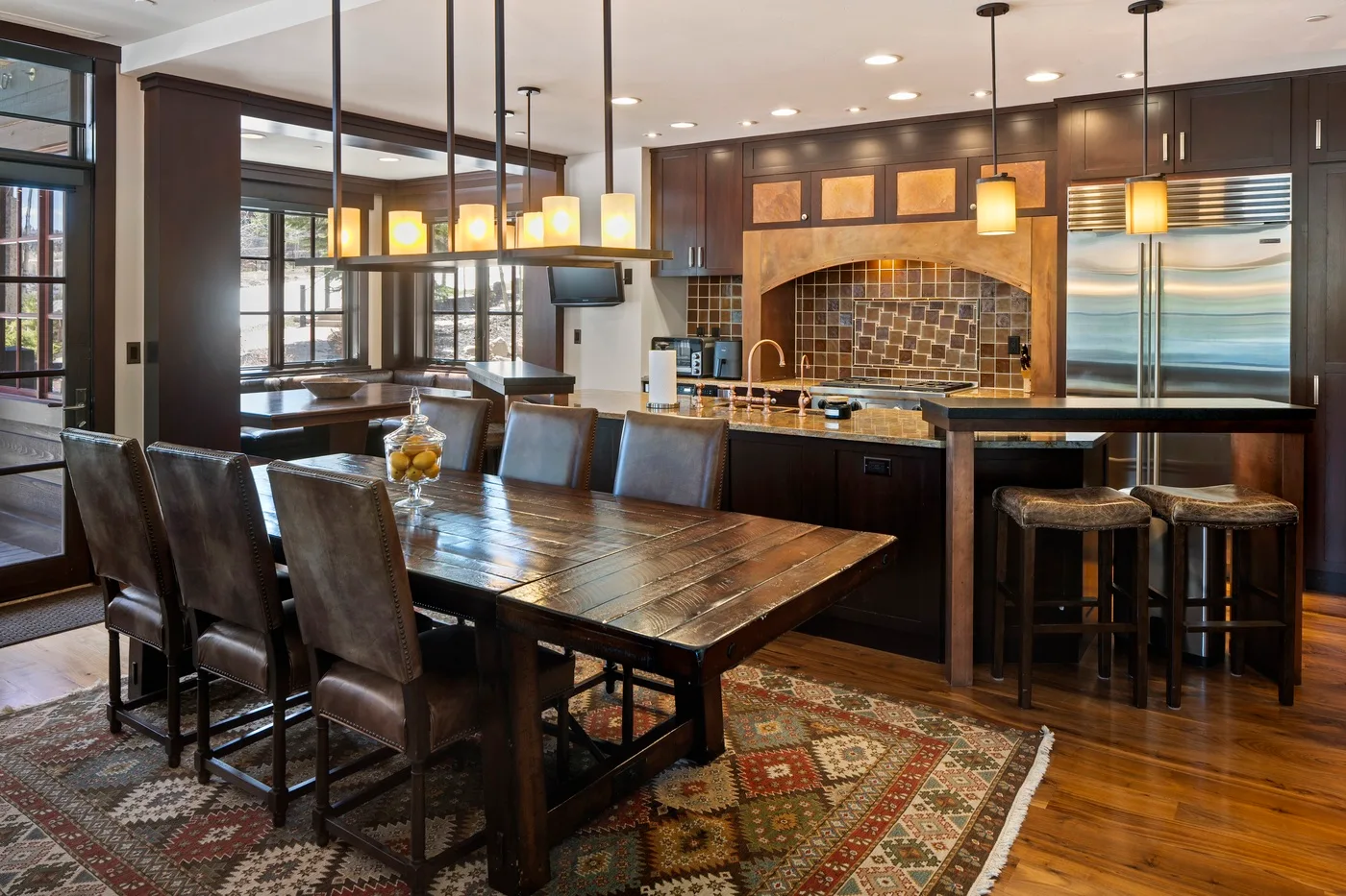A dining area beside a kitchen, with a long wooden table and hanging lights.