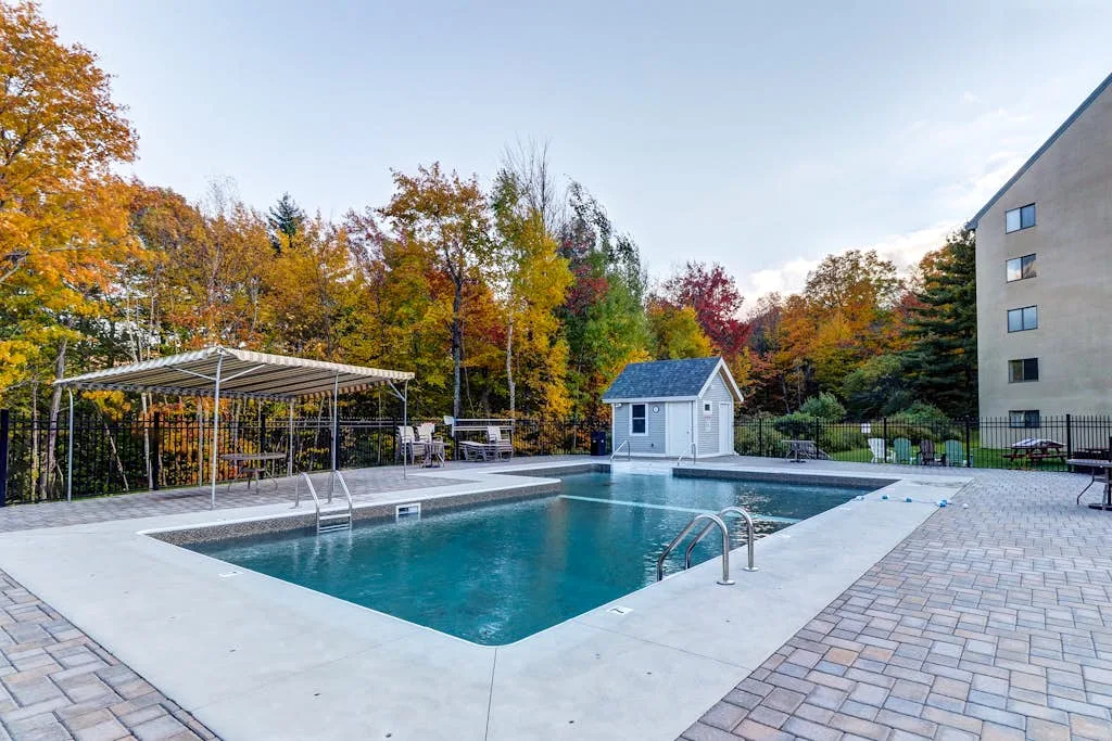 An outdoor swimming pool with a paved deck and autumn trees.