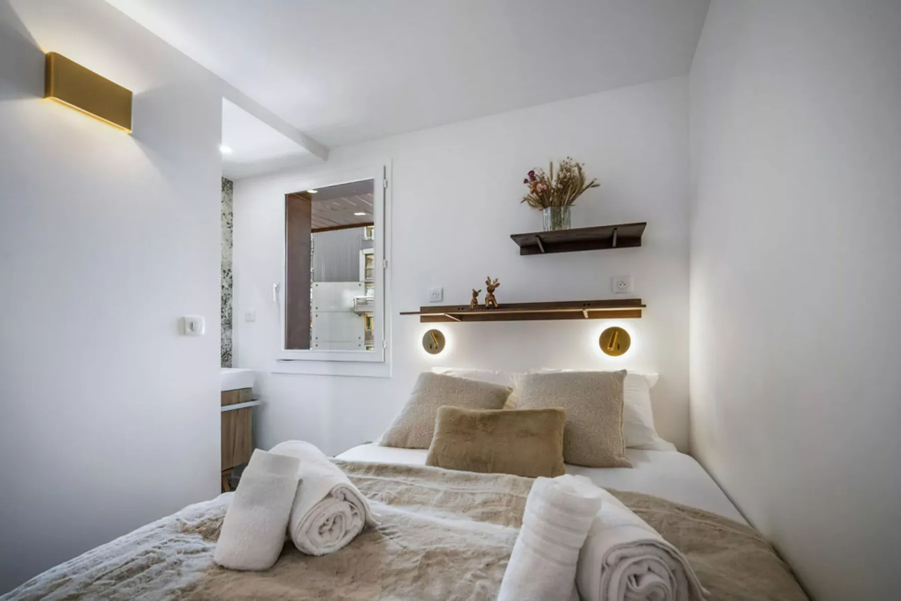 A bright bedroom with a bed, neutral bedding, and shelves on the wall.