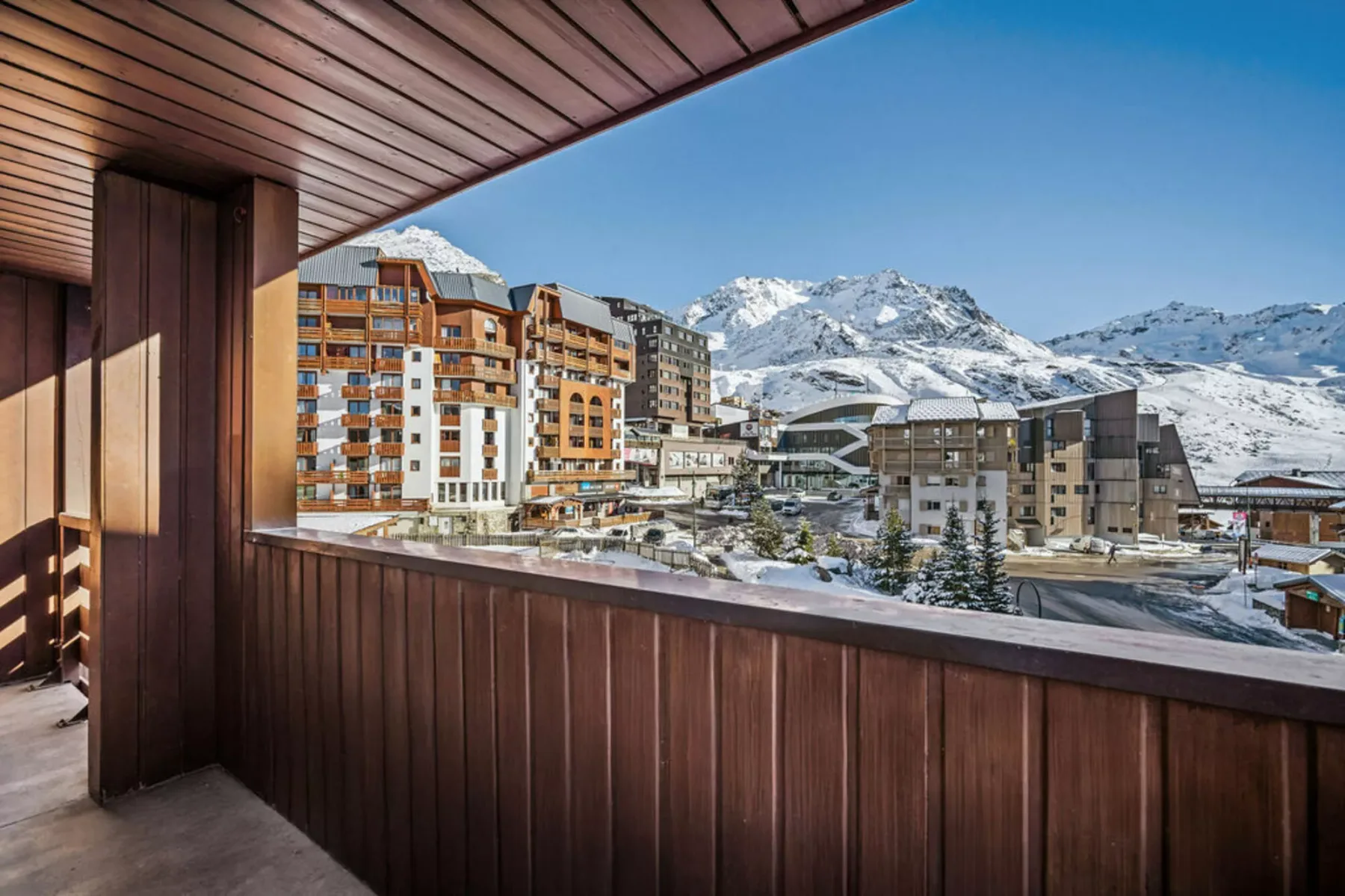 A balcony overlooks alpine buildings and snow-covered mountains beneath a clear blue sky.