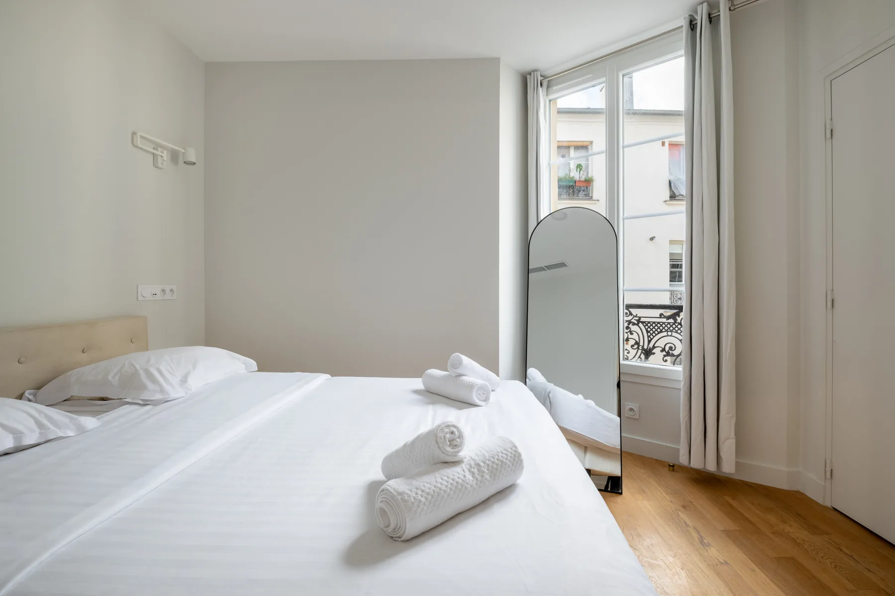 A bright bedroom with a neatly made bed and a tall mirror beside the window.