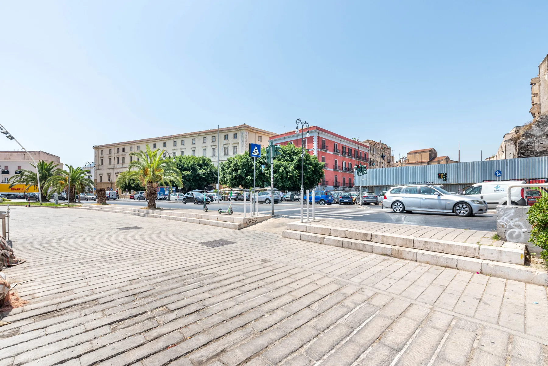 A paved city plaza with parked cars, palm trees, and multistory buildings.
