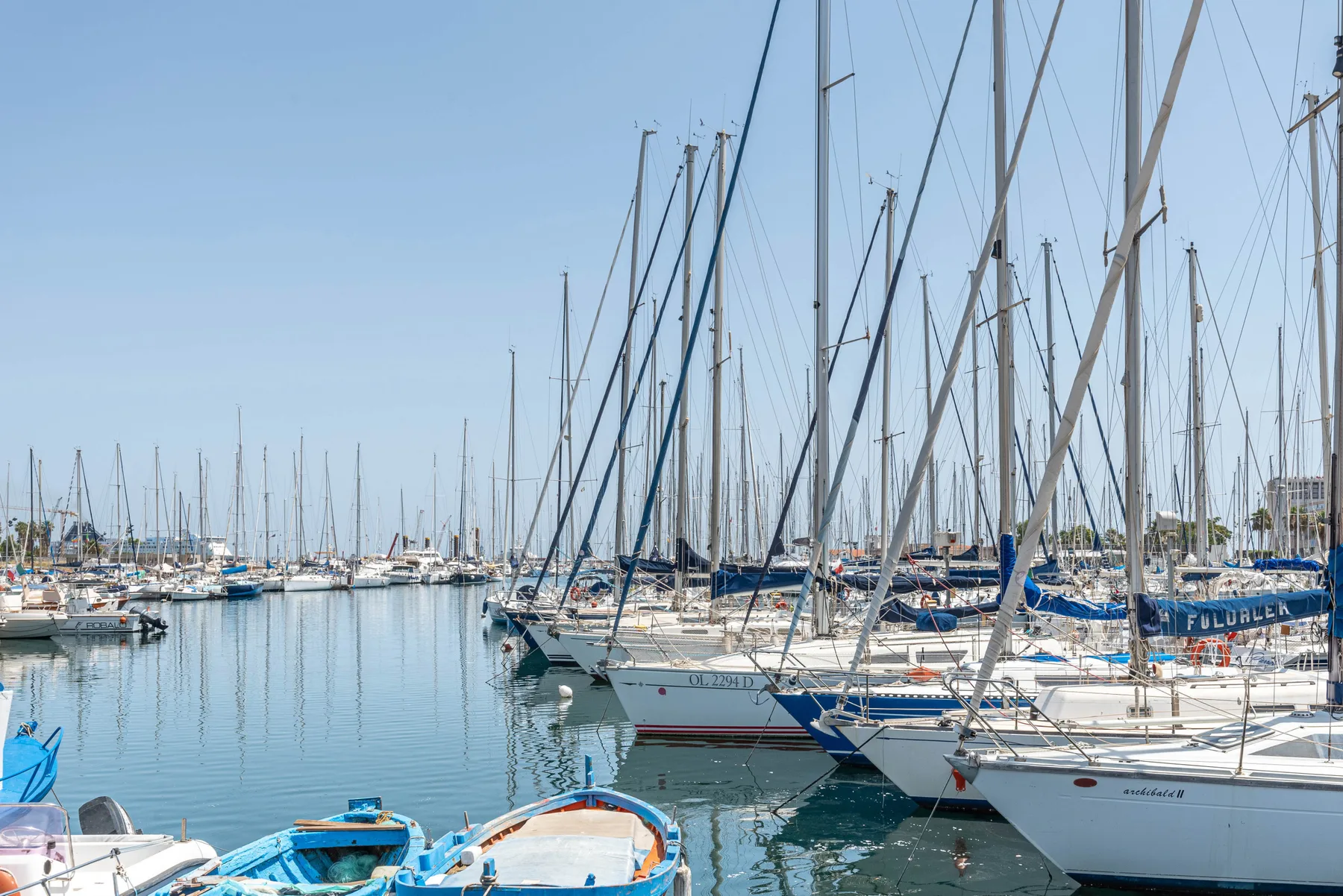A marina filled with sailboats sits along calm blue water under a clear sky.