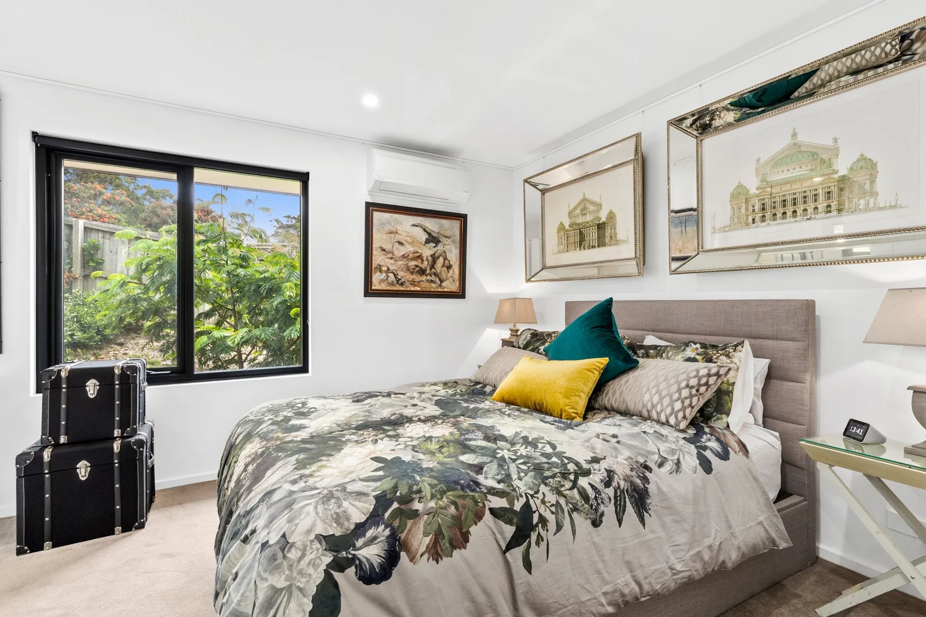 Un dormitorio con una colcha floral, cuadros enmarcados y una ventana con vistas a la vegetación.