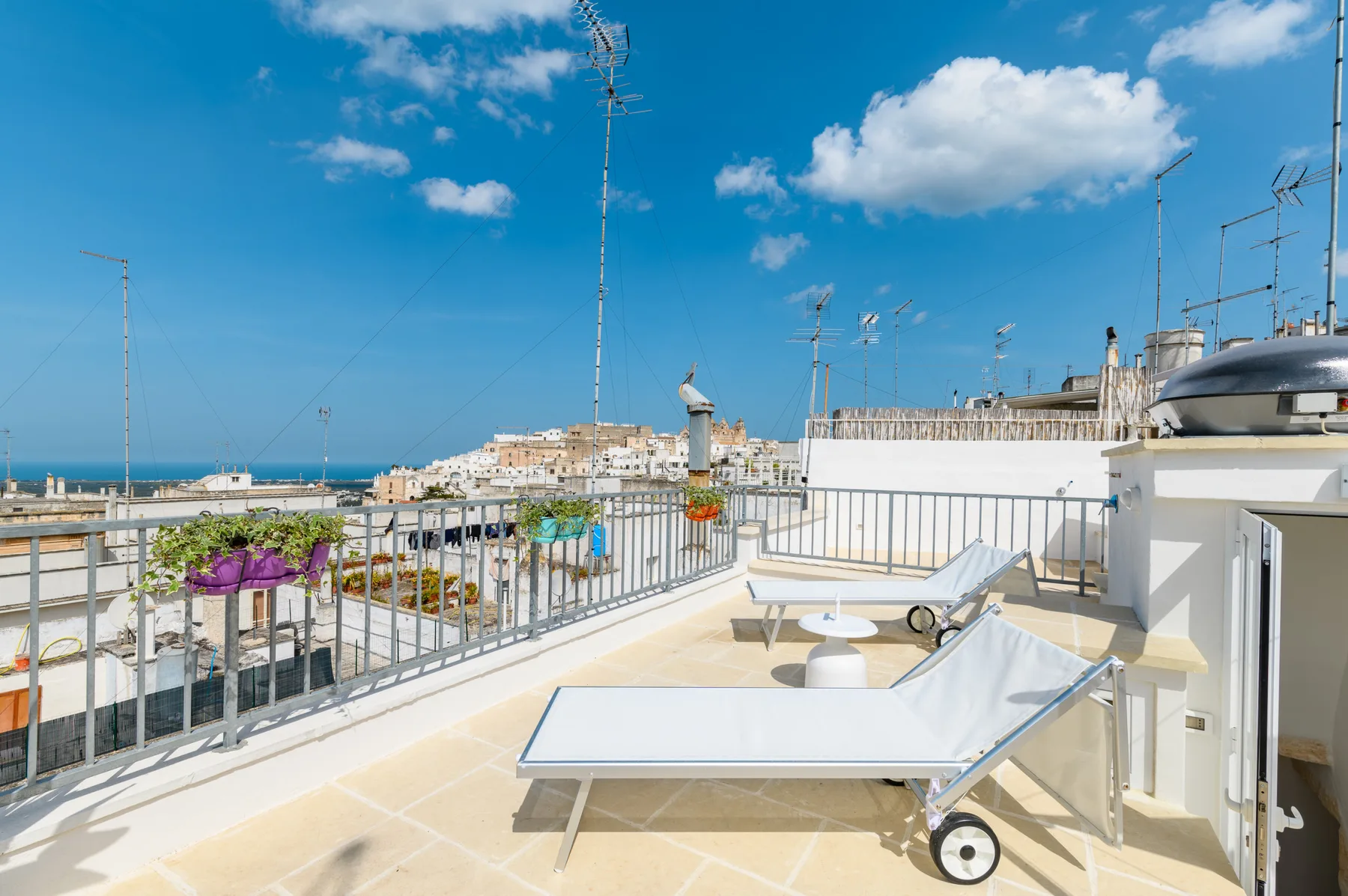 A sunny rooftop terrace has lounge seating, railings, and a view toward the sea.