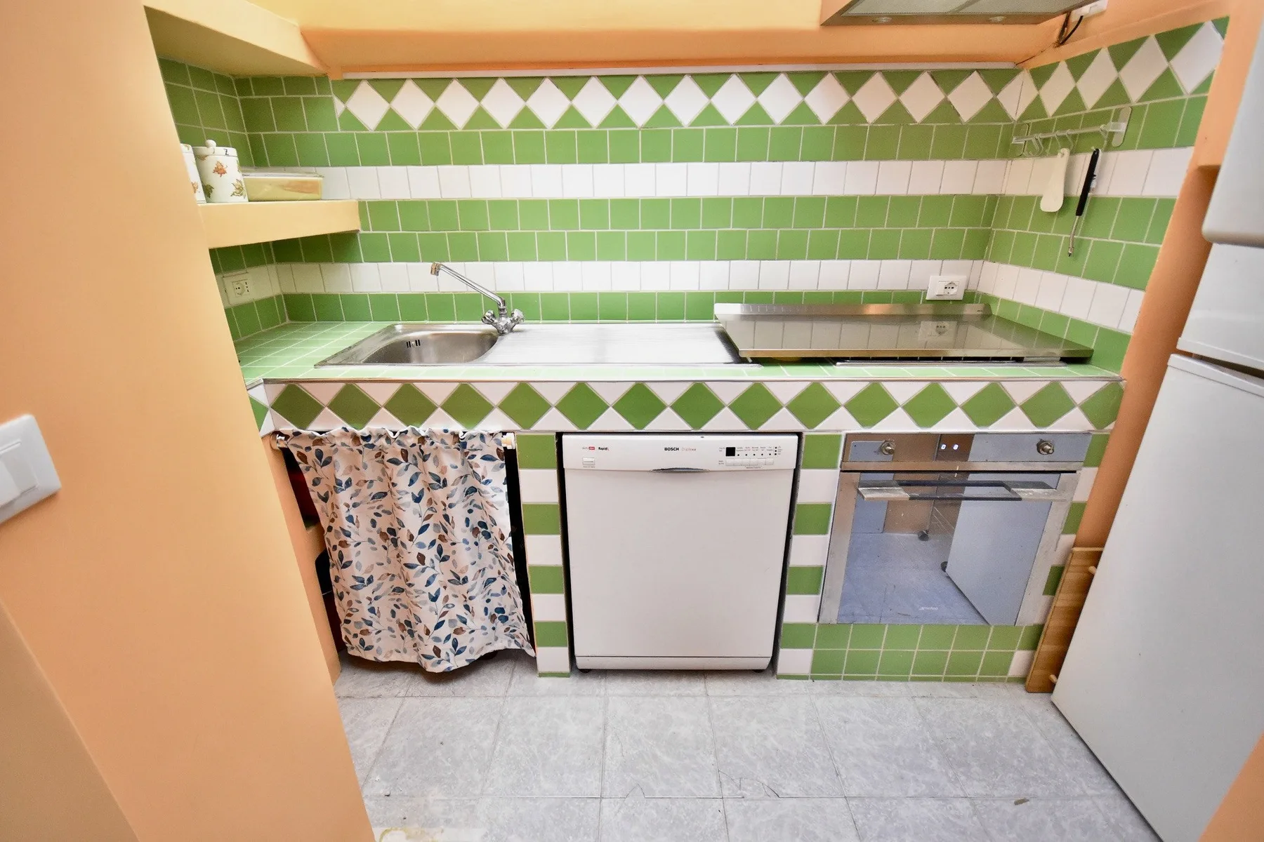 A compact kitchen with a green tiled counter, sink, cooktop, and built-in appliances.