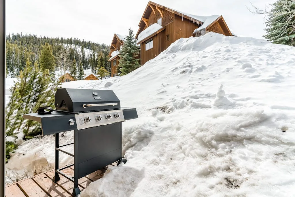 Una parrilla está sobre la nieve junto a una cabaña de montaña de madera.