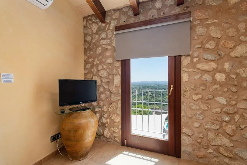 A stone-walled room has a glass door opening onto a balcony with a green landscape view.