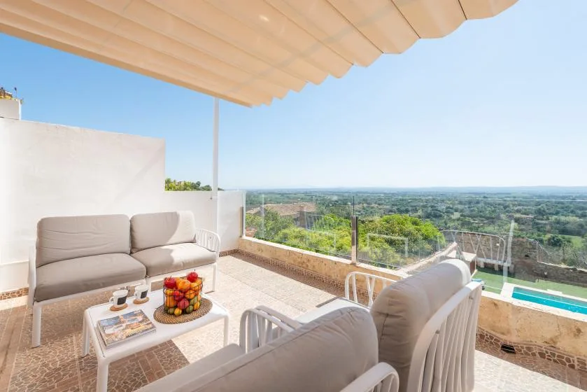 A covered terrace with seating overlooks a green landscape and swimming pool.