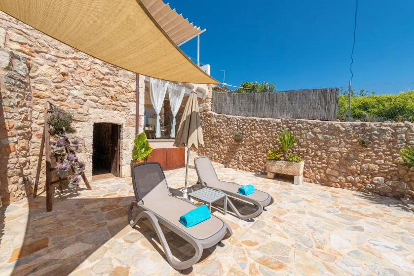 A sunlit stone patio with lounge seating, a shade canopy, and a low stone wall.
