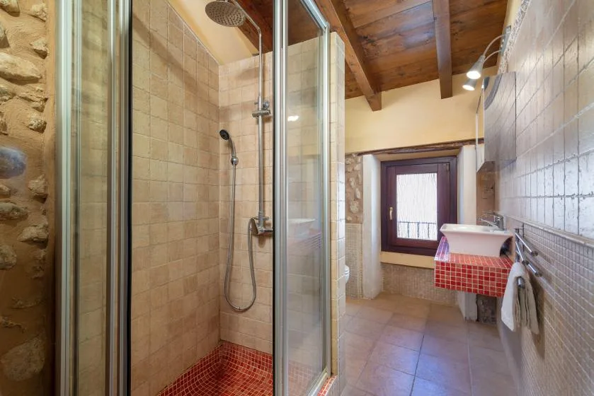 A tiled bathroom with a glass shower, sink, and window beneath a wooden ceiling.