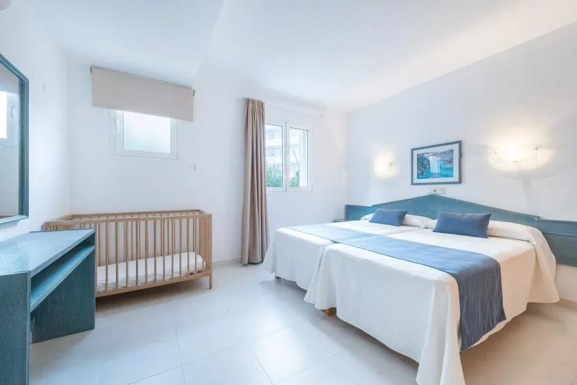 A bright bedroom with a double bed, a wooden crib, and a window.