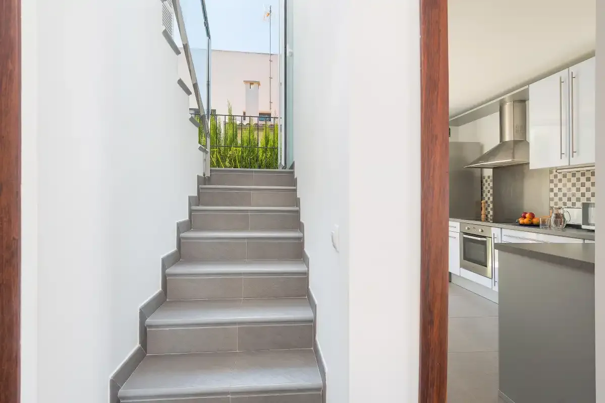Una escalera de baldosas sube junto a una cocina abierta con armarios blancos.