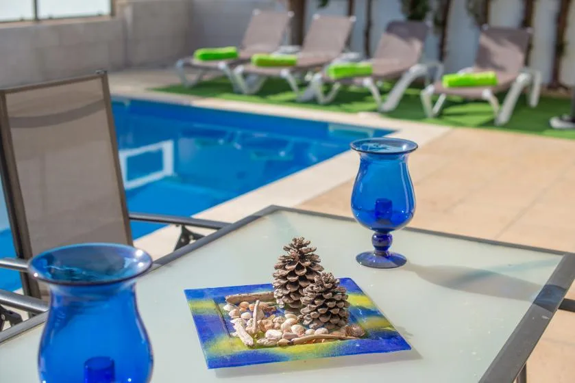 Una mesa de cristal en el patio con cristalería azul está junto a una piscina al aire libre.
