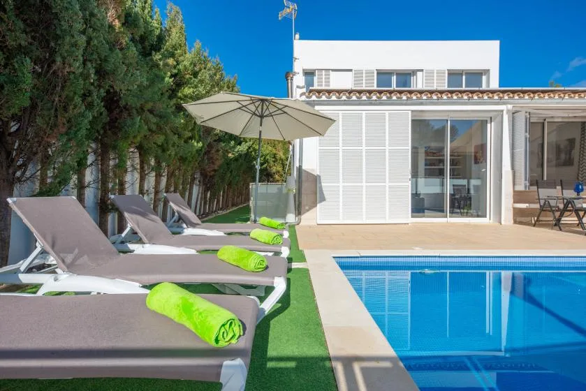 Una piscina al aire libre junto a una casa blanca moderna, con tumbonas junto al borde.