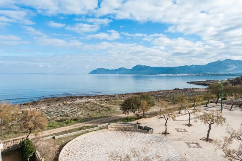 Paisaje costero con una terraza pavimentada, árboles dispersos y montañas al otro lado del agua.