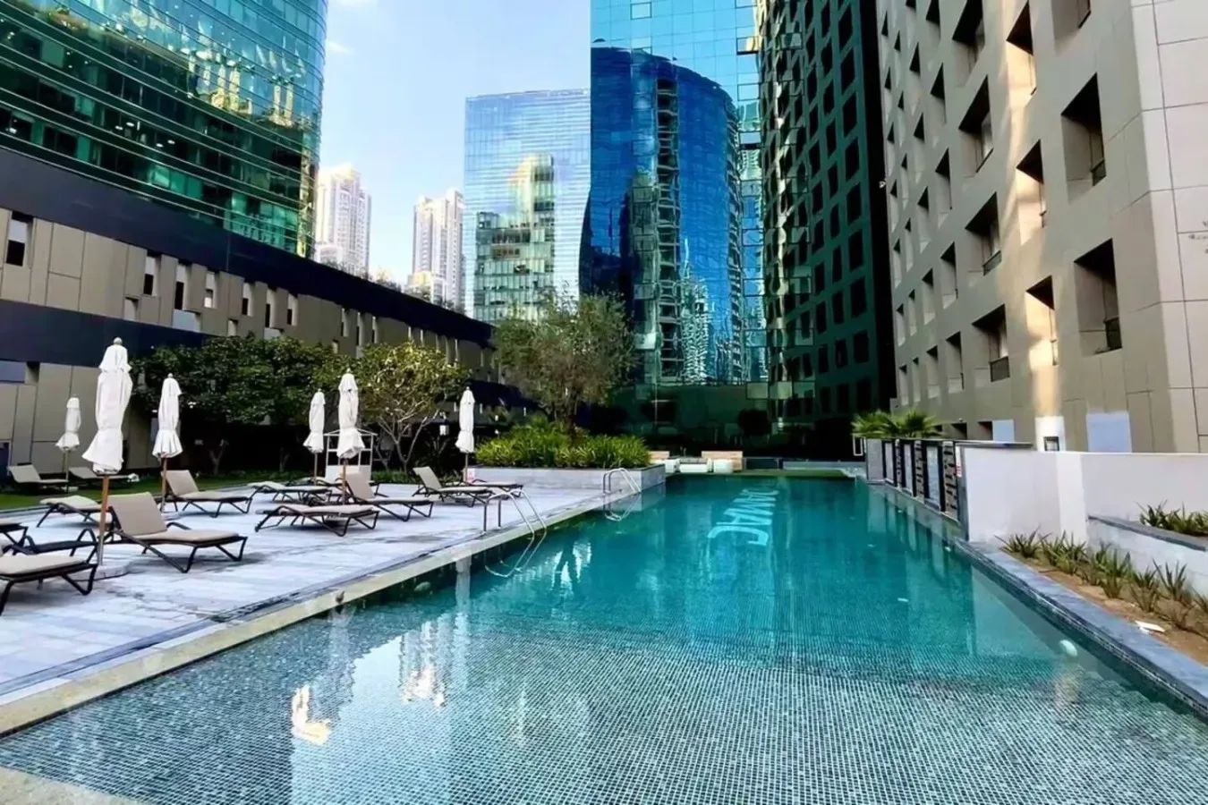 A swimming pool runs through a courtyard between modern buildings, with lounge seating along the edge.