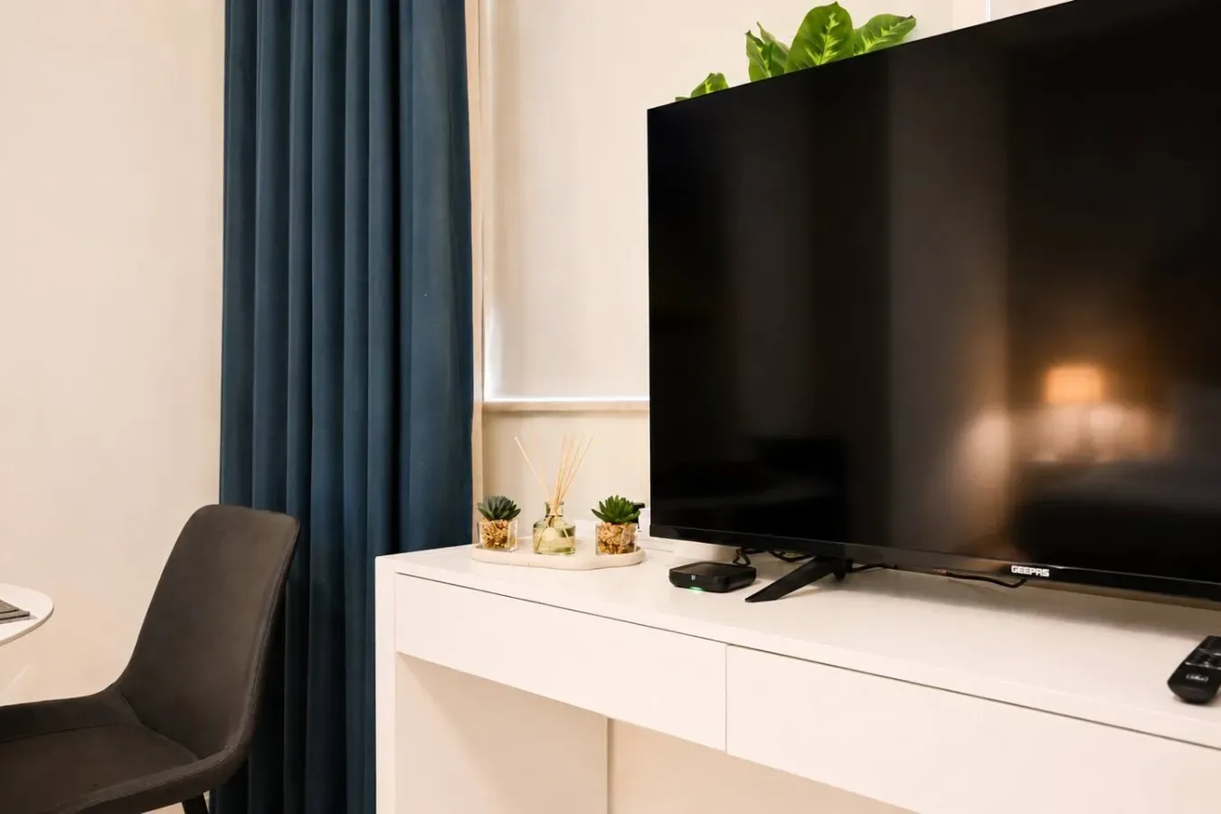 Un televisor está sobre una consola blanca junto a una cortina oscura y una silla.