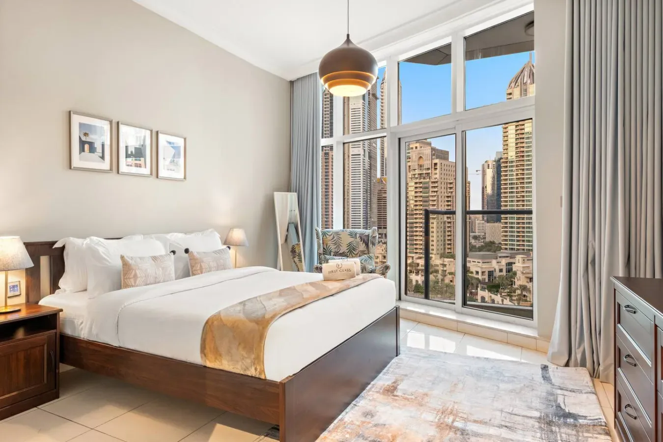 Un dormitorio luminoso con una cama bien arreglada junto a grandes ventanas con vistas a edificios de la ciudad.