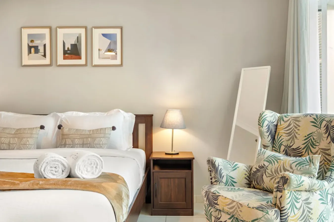 Dormitorio con una cama bien tendida y un sillón estampado junto a la ventana.