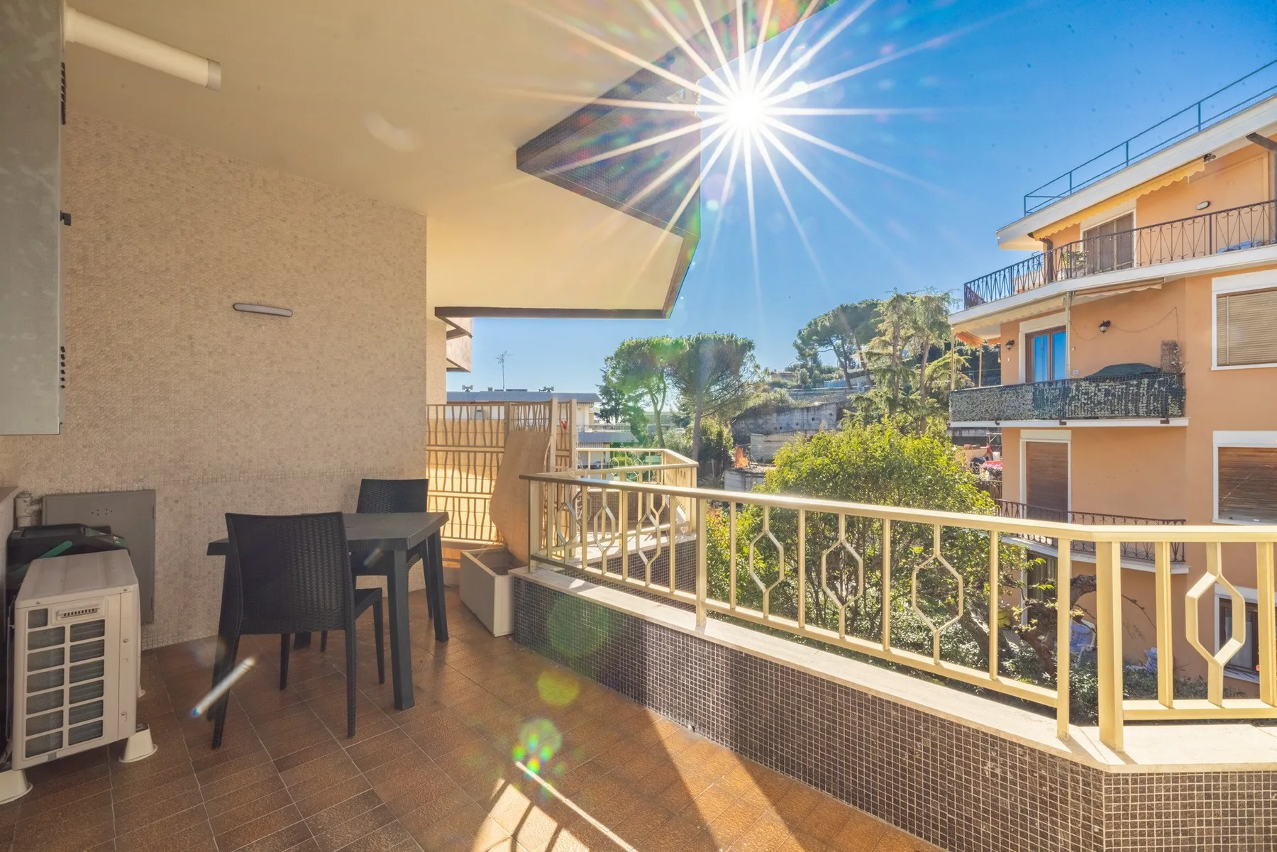 A sunlit balcony with outdoor seating and a railing overlooking nearby buildings and trees.