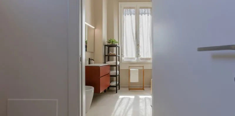 A bright bathroom with a vanity, shelving, and a curtained window.
