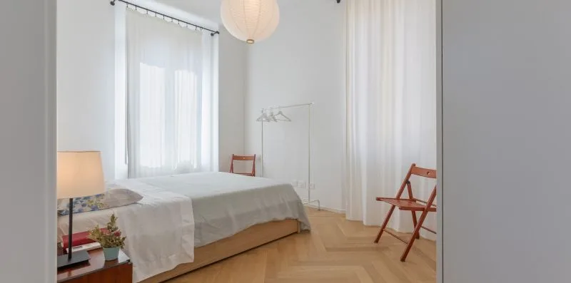 A bright bedroom with a bed, sheer curtains, and a folding chair.