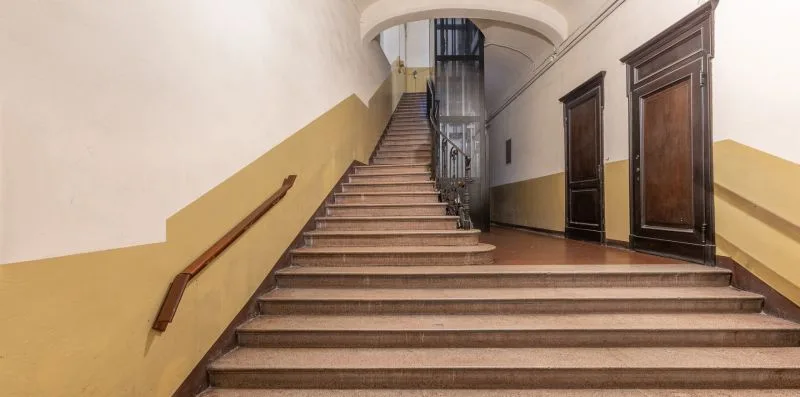 A broad indoor staircase rises beside dark wooden doors and a metal railing.