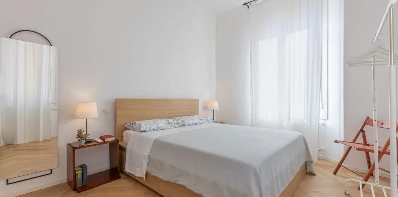 A bright bedroom with a double bed, bedside table, and sheer curtains.