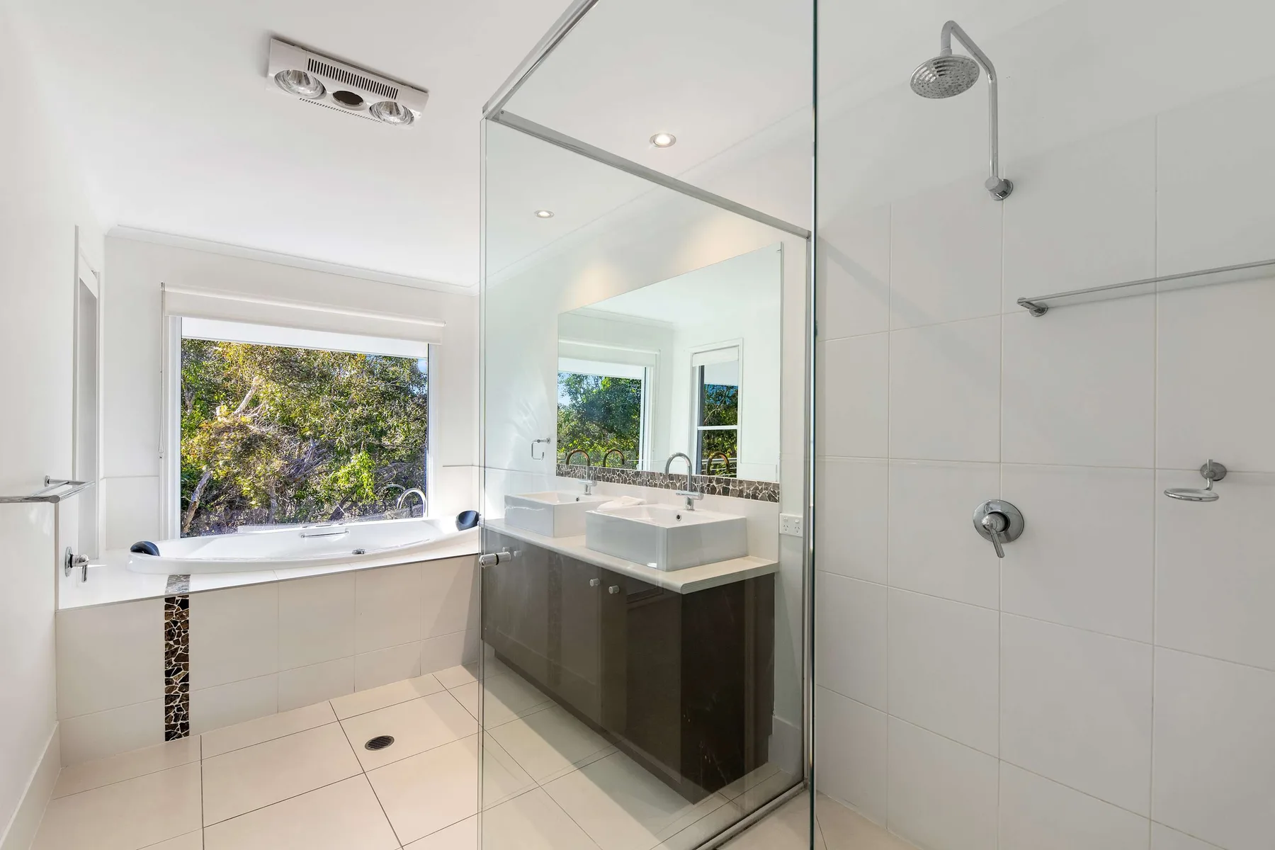 A bright bathroom has a glass shower, vanity, and window overlooking greenery.