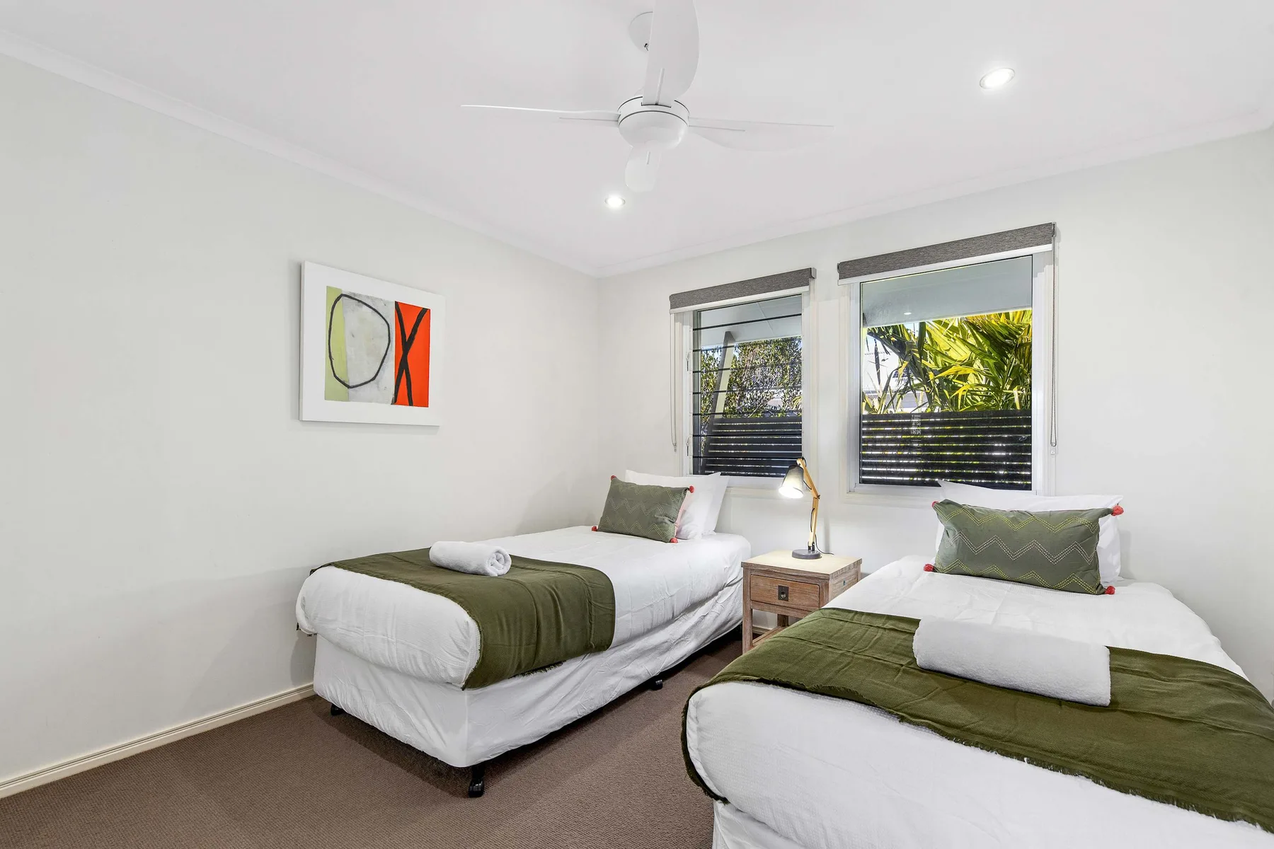 A bright bedroom with two beds and windows overlooking greenery.