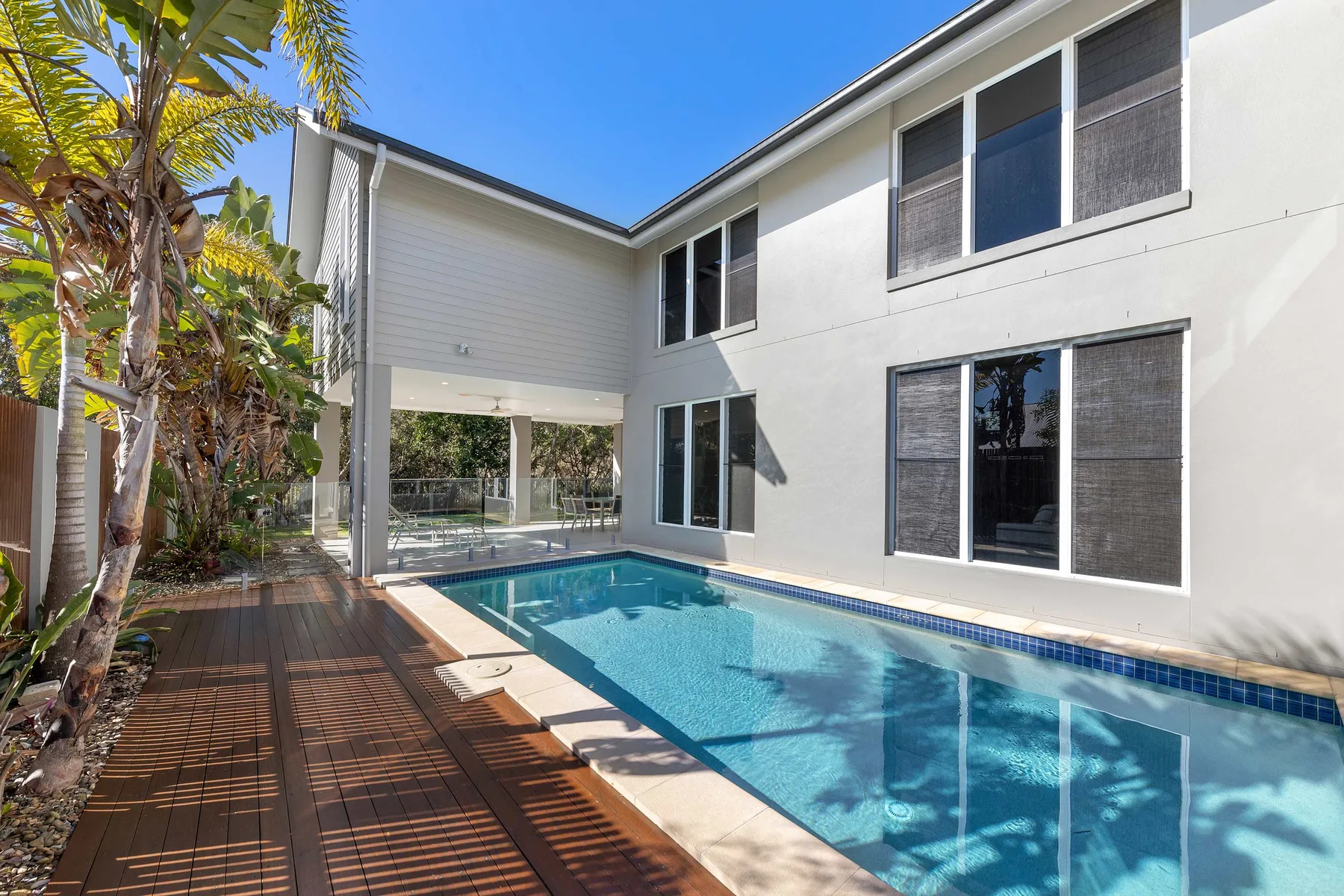 A swimming pool beside a modern house, with a wooden deck and tropical plants.