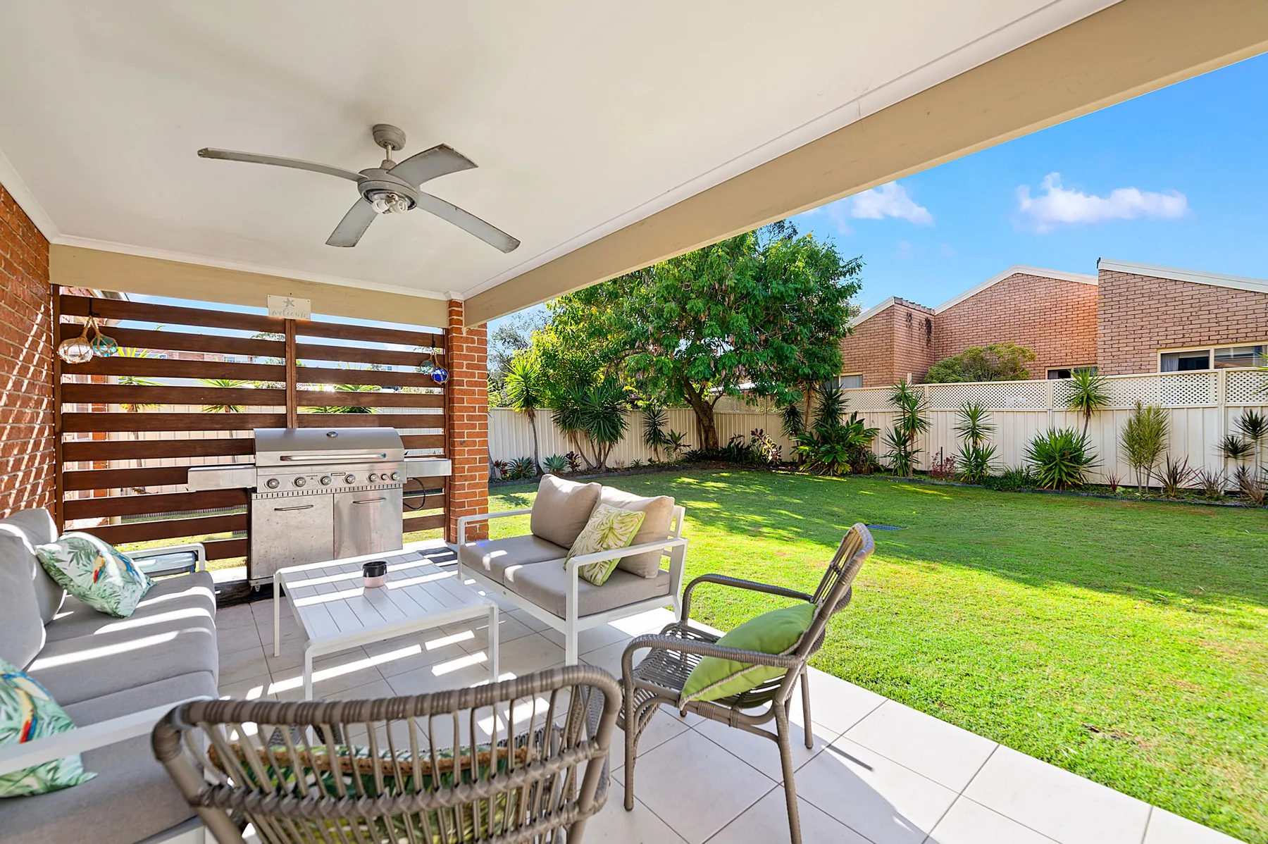 A covered outdoor patio has seating beside a barbecue, overlooking a green lawn.