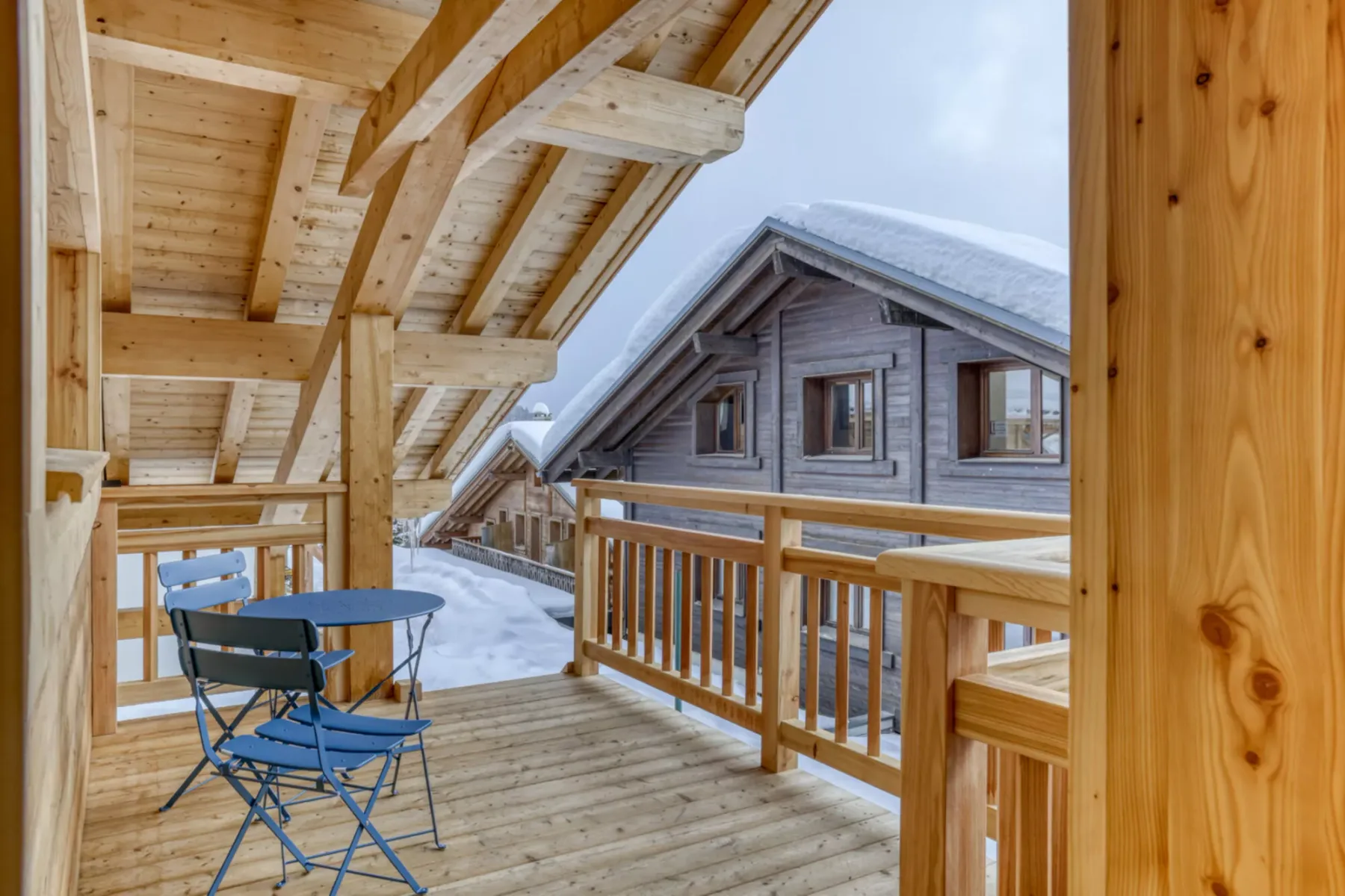 A covered wooden balcony overlooks a snow-covered chalet and outdoor seating.
