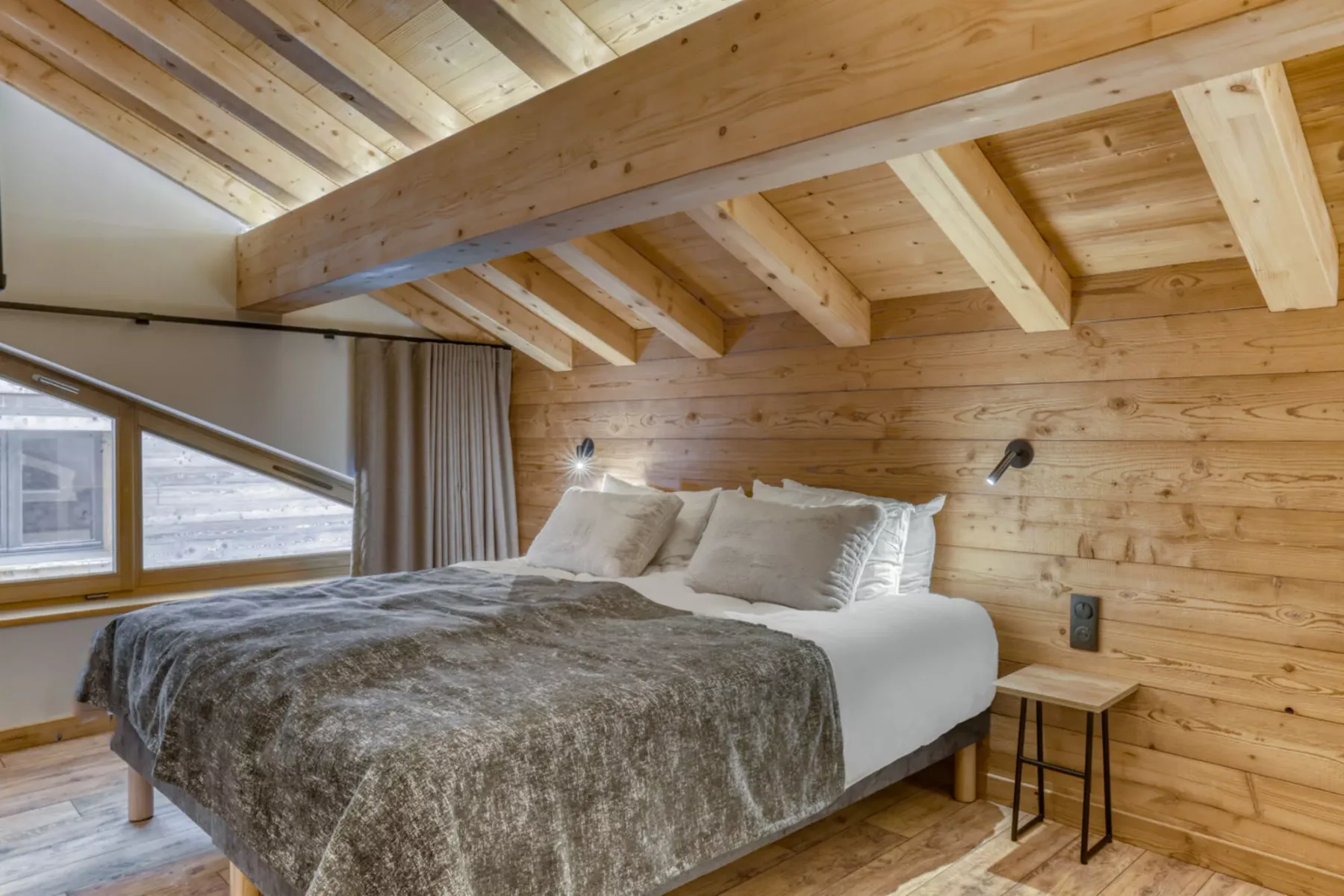 A bedroom with a bed beneath exposed wooden ceiling beams and beside windows.