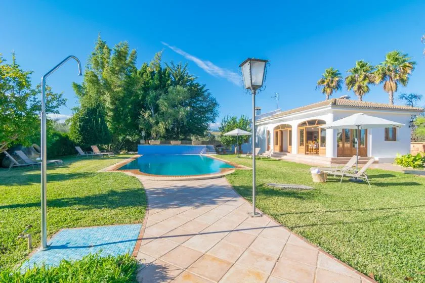Una piscina en el jardín junto a una casa de una planta, con camino pavimentado y césped.