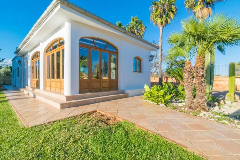 Un edificio blanco con ventanas arqueadas enmarcadas en madera, junto a un patio embaldosado y palmeras.