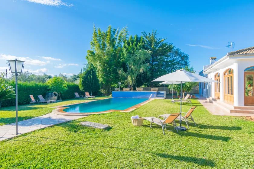 Un jardín soleado rodea una piscina, con asientos al aire libre junto a una casa blanca.