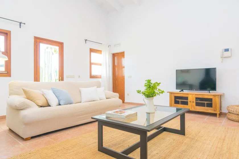 Una sala de estar luminosa con asientos beige, una mesa de centro de vidrio y un televisor.