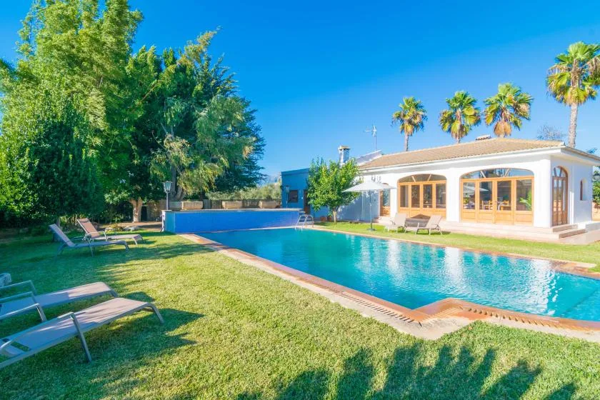 Una piscina junto a una casa luminosa, con asientos de descanso en el césped.
